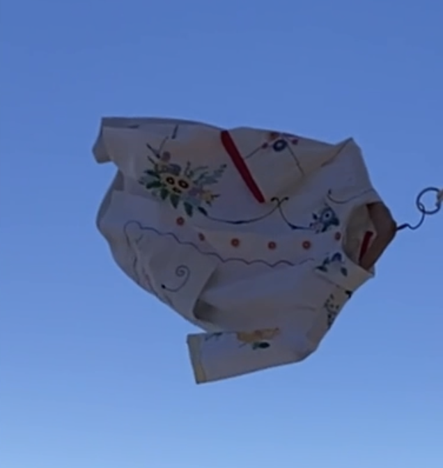 short-sleeved shirt made from recycled tablecloths is mainly white and off-white with colourful embroidered and appliquéd flowers and orange buttons, shown flying through the air on a windy day