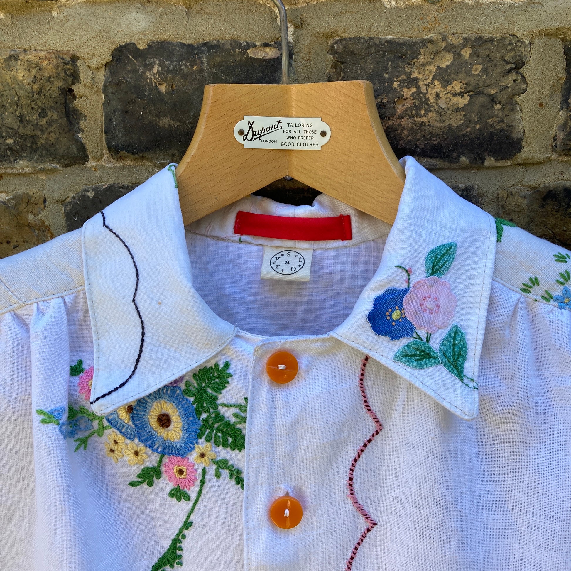 Close up of collar of short-sleeved shirt made from recycled tablecloths is mainly white and off-white with colourful embroidered and appliquéd flowers and orange buttons