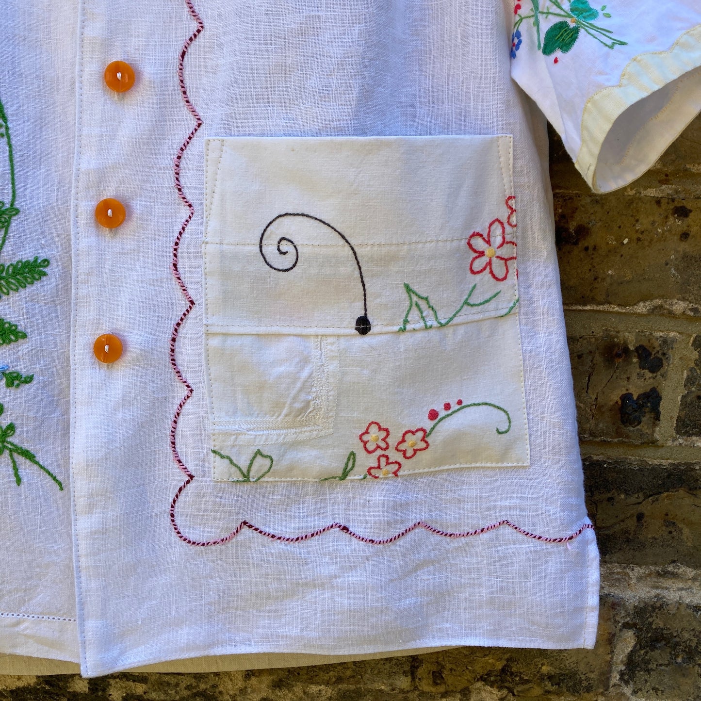 Close-up of pocket with original visible mending on short-sleeved shirt made from recycled tablecloths is mainly white and off-white with colourful embroidered and appliquéd flowers and orange buttons