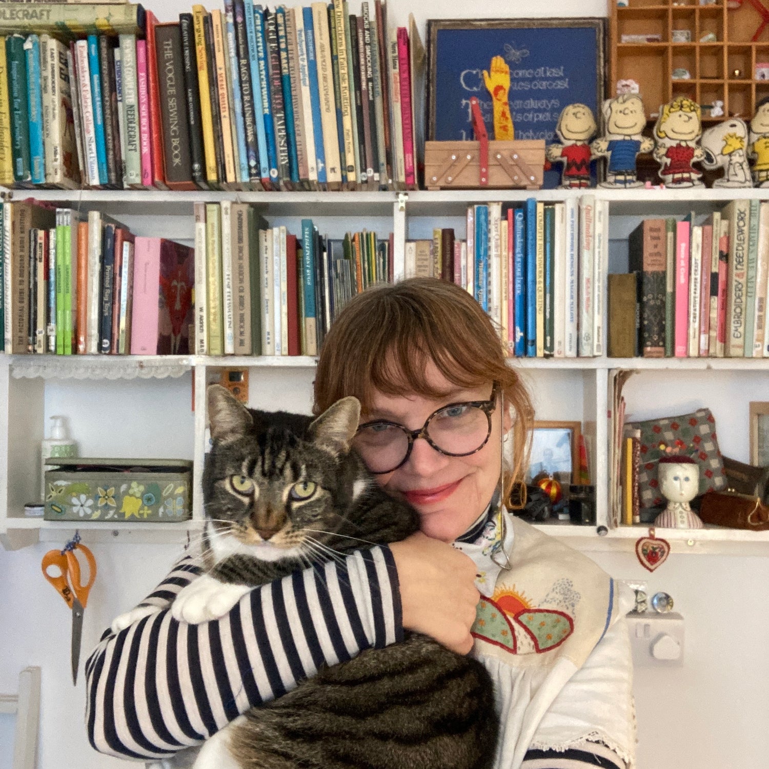 Elizabeth Cake, a white woman with auburn hair and glasses, the designer and maker behind A Circular Story stands in front of some bookshelves holding her tabby cat, Cloudfeet