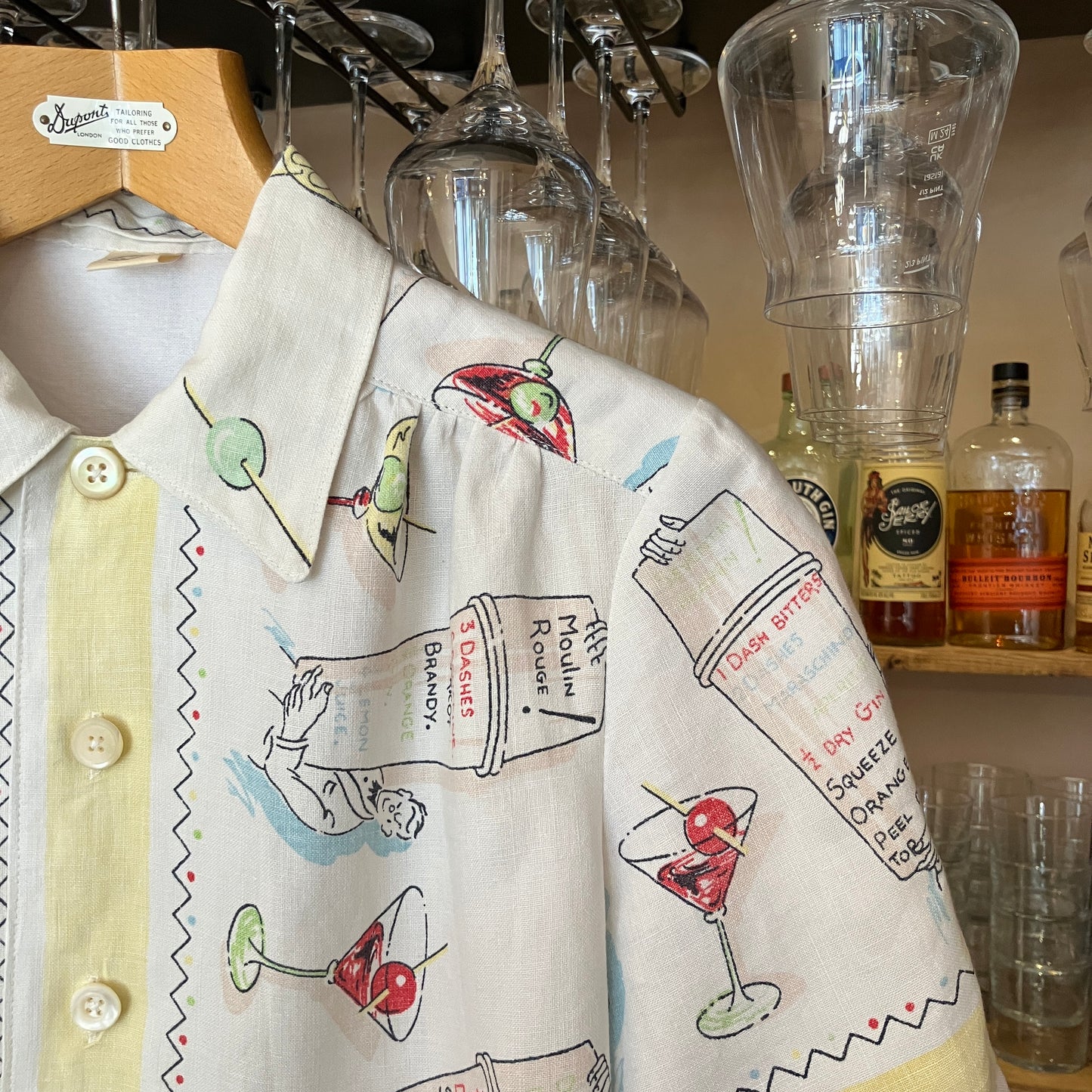 Shirt with cocktail-themed design hanging next to bottles and glasses in a bar setting