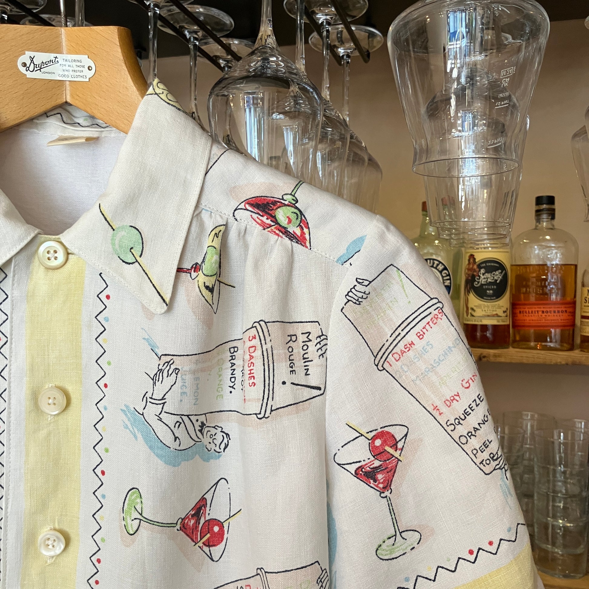 Shirt with cocktail-themed design hanging next to bottles and glasses in a bar setting