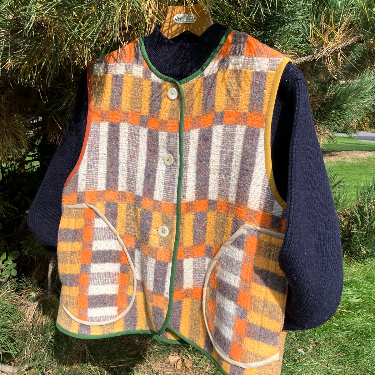 A vintage patterned wool vest with orange, yellow, gray, and white plaid panels, featuring green trim and button closure, layered over a navy blue knit sweater. The vest is displayed on a wooden hanger hanging from pine tree branches outdoors.