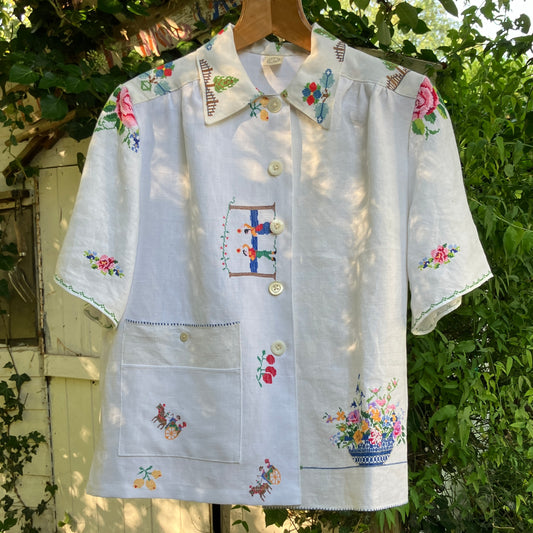 Linen shirt made from a patchwork of recycled linen tablecloths with cross-stitch motifs including dancers, flowers, trees, lemons, tomatoes and people in a horse-drawn chariot