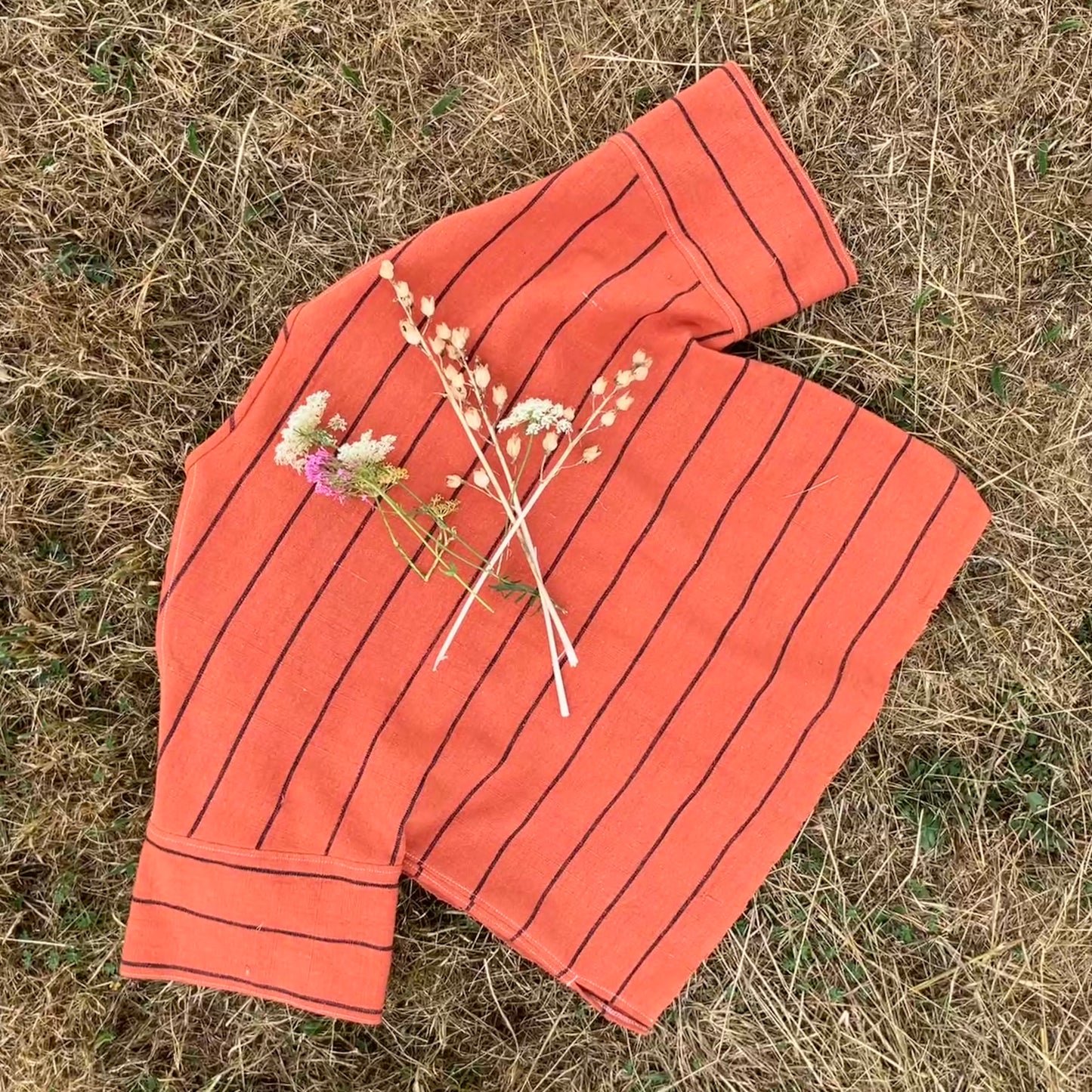 A terracotta orange boxy top with thin black stripes, laid flat on dry grass surrounded by dried flowers 