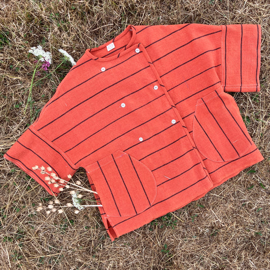 A terracotta orange boxy top with thin black stripes, laid flat on dry grass surrounded by dried flowers 