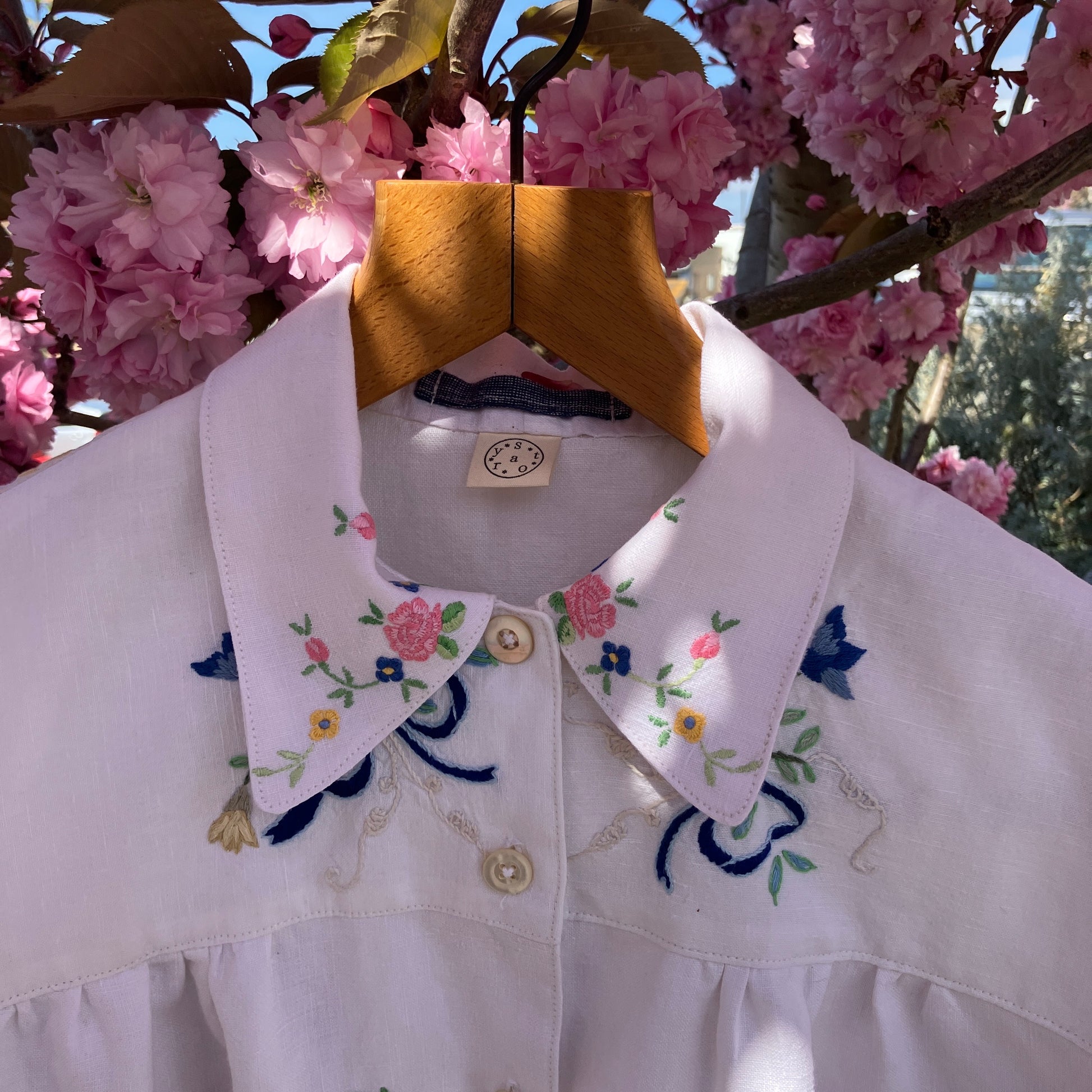 Short sleeved shirt made from a patchwork of reclaimed tablecloths with a traditional English pub with a thatched roof embroidered on the front and two bows on the yoke, close-up, hanging in a pink blossom tree