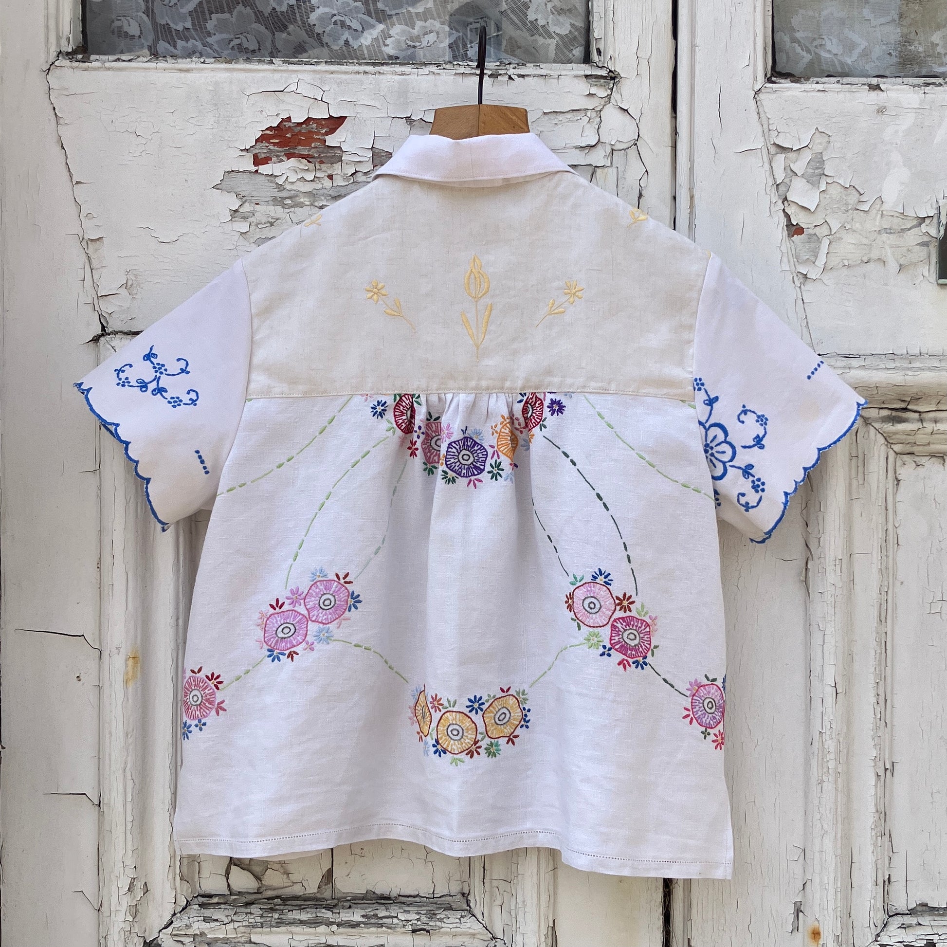 Short sleeved shirt made from a patchwork of reclaimed tablecloths with a traditional English pub with a thatched roof embroidered on the front and two bows on the yoke (bank view)