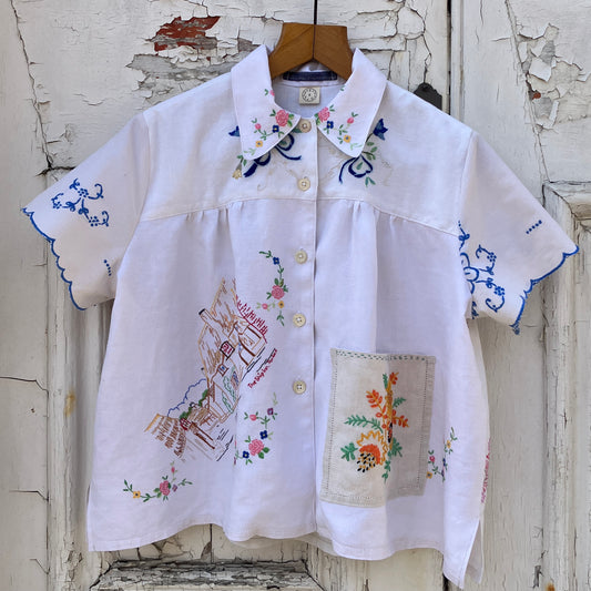 Short sleeved shirt made from a patchwork of reclaimed tablecloths with a traditional English pub with a thatched roof embroidered on the front and two bows on the yoke