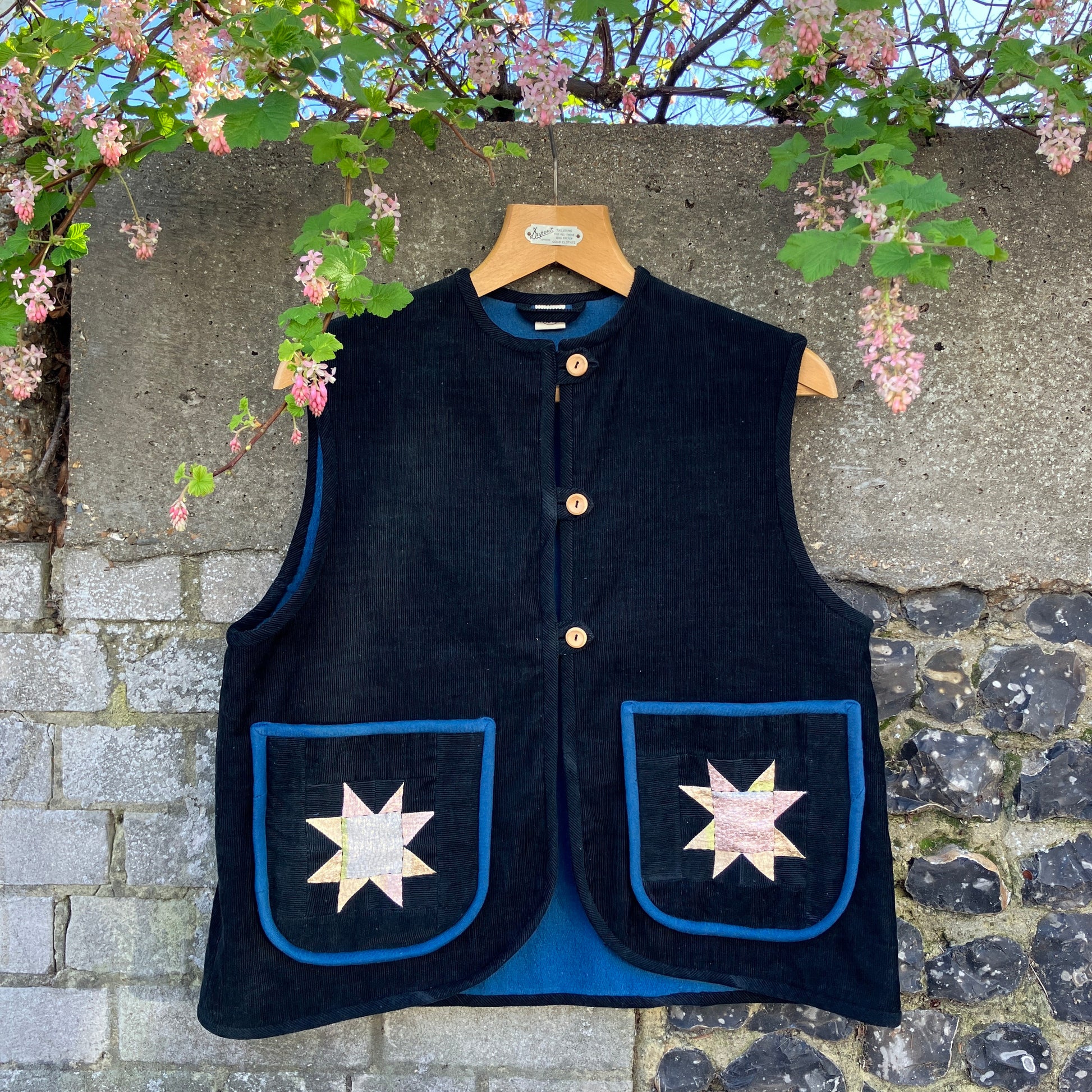 Needlecord vest/gilet/waistcoat with large hand quilted patchwork pockets in gold and silver lamé with blue binding.  Finished with wooden buttons and a hanging loop. Pictured hanging against a wall with spring blossom overhanging