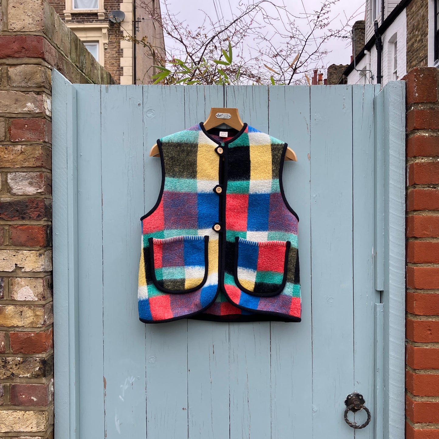 A vest/gilet/waistcoat made from a multicoloured checked reclaimed blanket. It fastens with three wooden buttons and has to large patch pockets