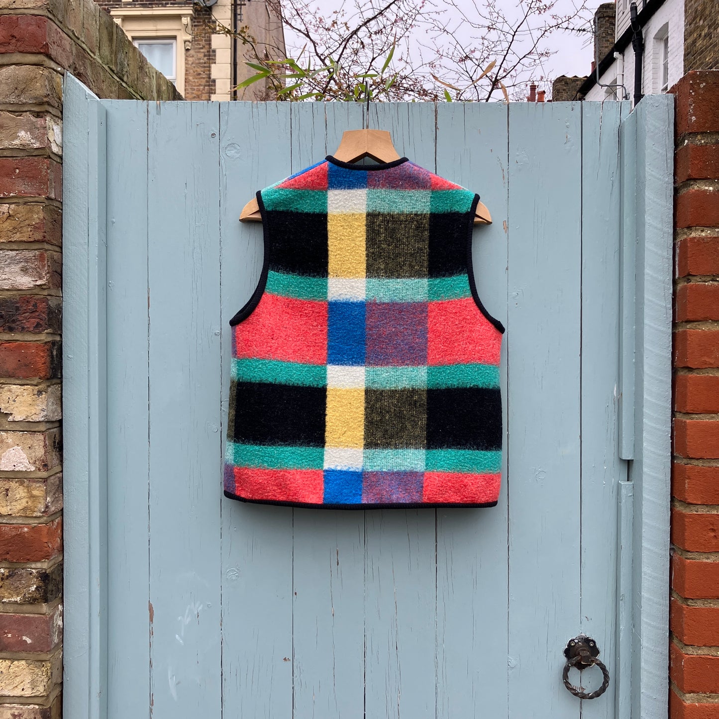 A vest/gilet/waistcoat made from a multicoloured checked reclaimed blanket. It fastens with three wooden buttons and has to large patch pockets (back view, hanging on a pale blue gate)