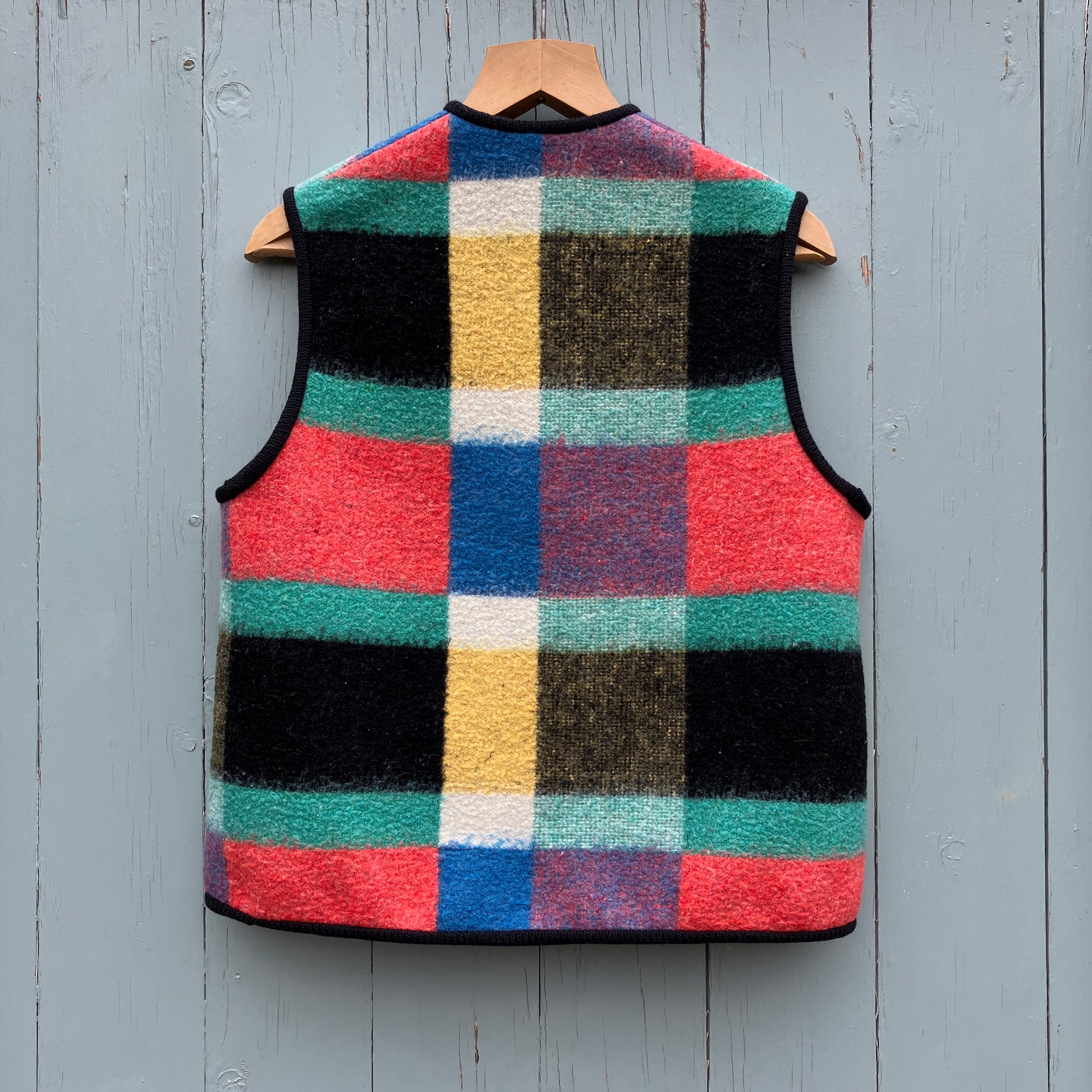 A vest/gilet/waistcoat made from a multicoloured checked reclaimed blanket. It fastens with three wooden buttons and has to large patch pockets (back view)