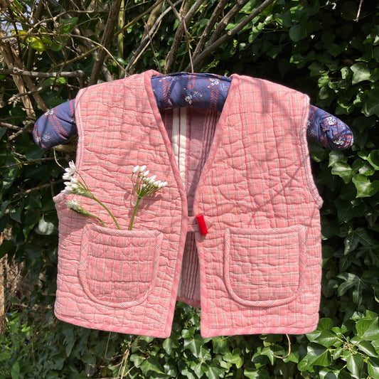 kids vest/gilet/waistcoat made from offcuts of a reclaimed pink quilt. Shown hanging on a blue vintage hanger in some foliage with some wild flowers in one of the pockets