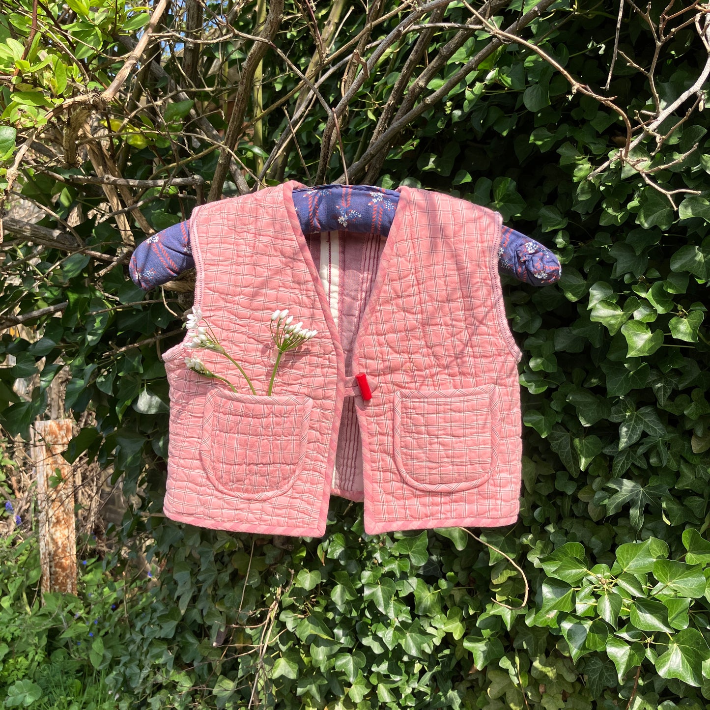 kids vest/gilet/waistcoat made from offcuts of a reclaimed pink quilt. Shown hanging on a blue vintage hanger in some foliage with some wild flowers in one of the pockets