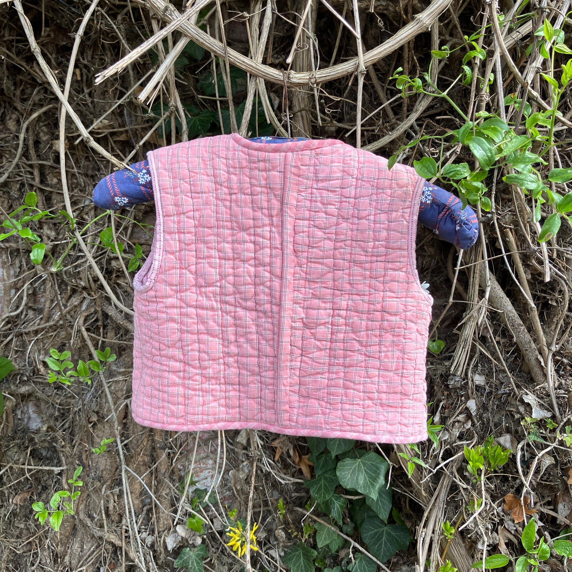 kids vest/gilet/waistcoat made from offcuts of a reclaimed pink quilt. Shown hanging on a blue vintage hanger in some foliage with some wild flowers in one of the pockets (back view)