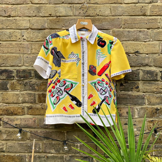 short-sleeved shirt made from a recycled tablecloth printed with picnic food a record player and records, mainly yellow and white, hanging on a hanger against a brick wall