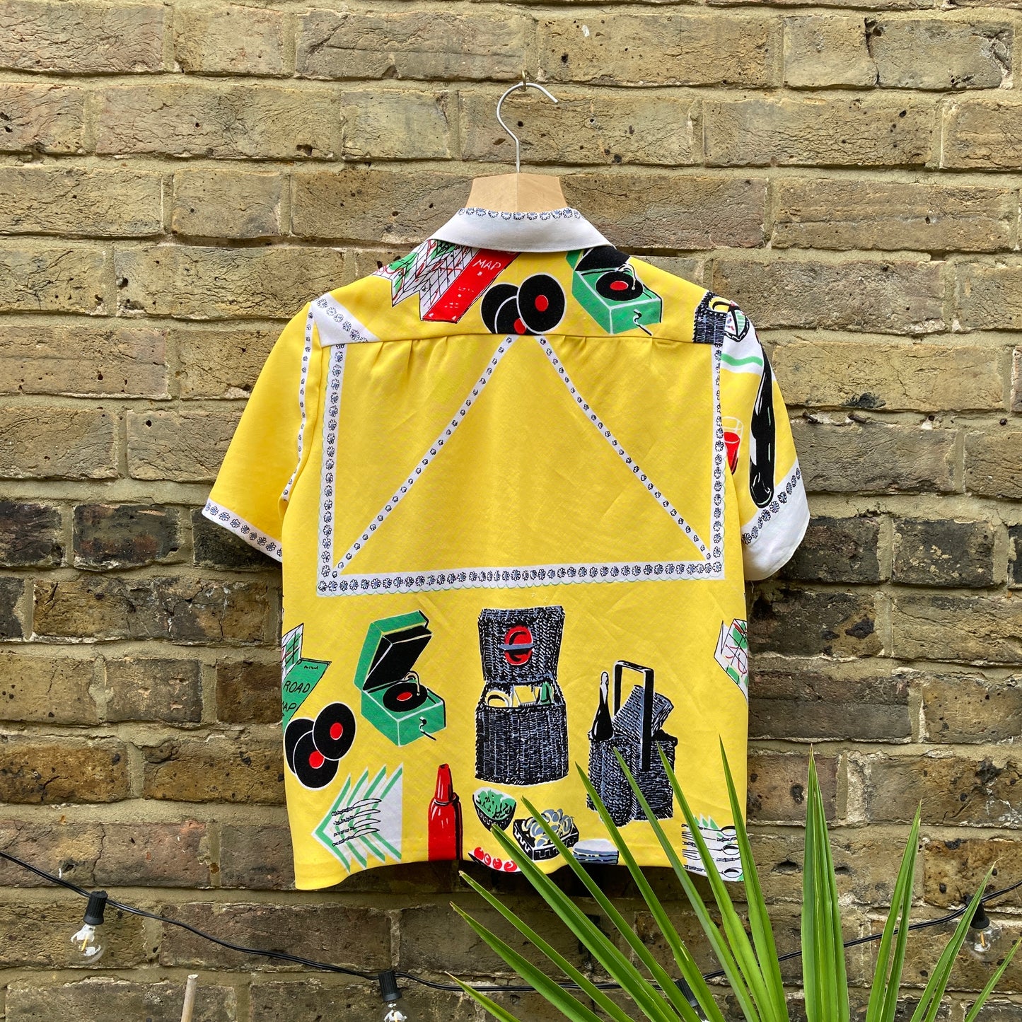 short-sleeved shirt made from a recycled tablecloth printed with picnic food a record player and records, mainly yellow and white, hanging on a hanger against a brick wall (back view)