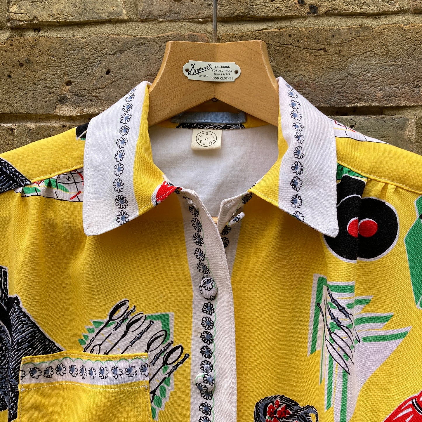 short-sleeved shirt made from a recycled tablecloth printed with picnic food a record player and records, mainly yellow and white, hanging on a hanger against a brick wall (detail of collar)