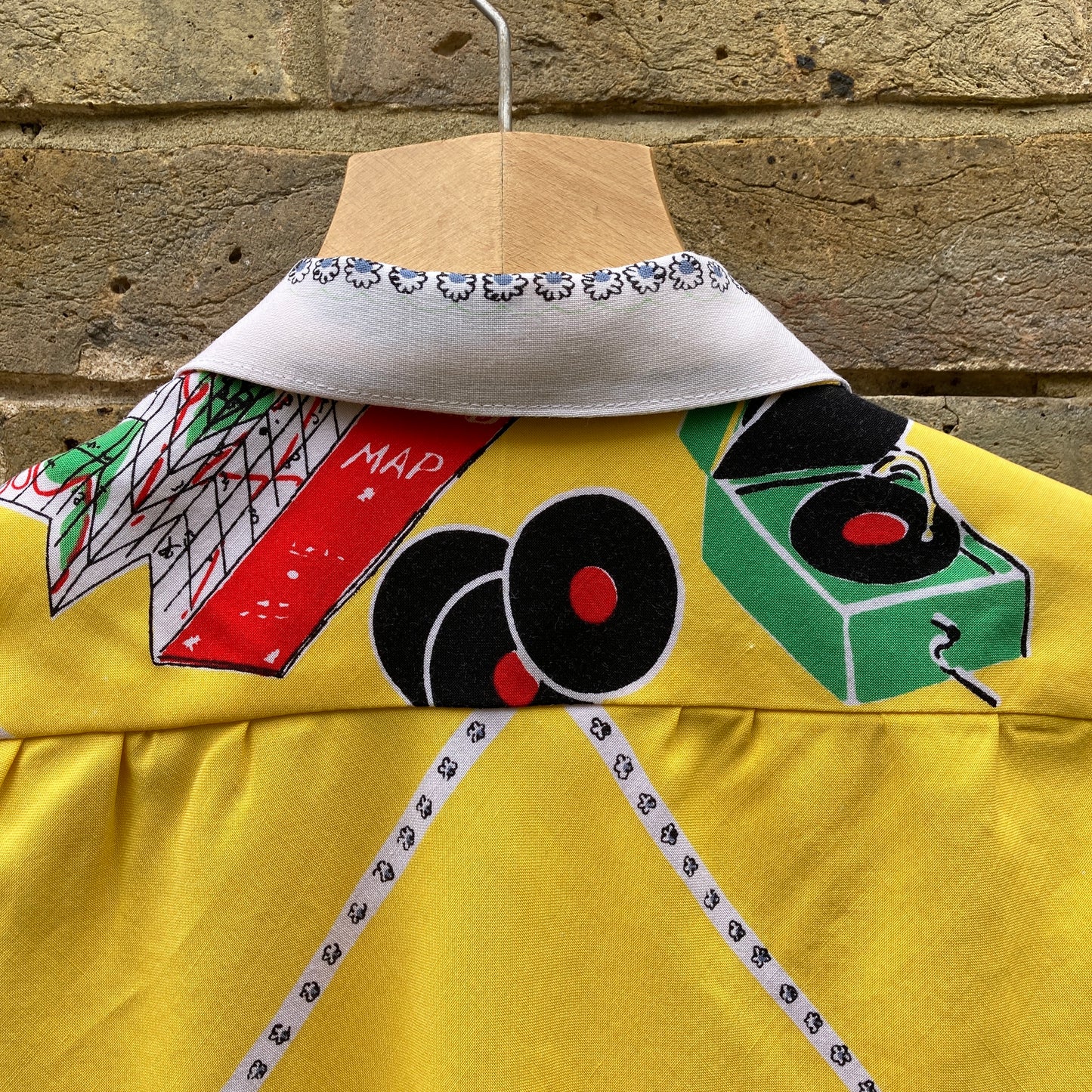 short-sleeved shirt made from a recycled tablecloth printed with picnic food a record player and records, mainly yellow and white, hanging on a hanger against a brick wall (close-up of back yoke)