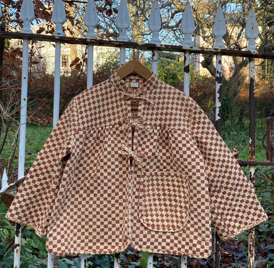 A quilted jacket in two shades of brown checkerboard print that fastens with three bows down the front. It has a big pocket and its hanging on an ornate but rusty gate