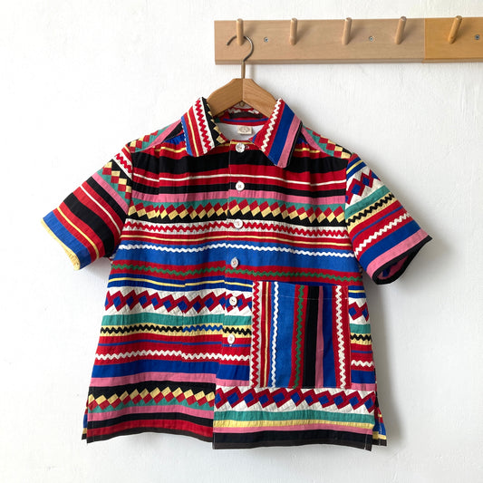 A short-sleeved shirt made from brightly coloured seminole patchwork hanging on a hanger from some wooden hooks