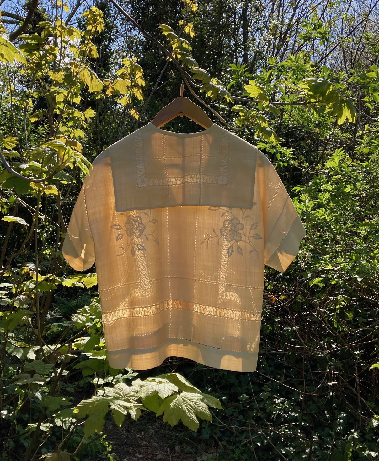 Sailor-collar shirt hand sewn from a reclaimed silk tablecloth, based on a vintage 80s pattern. Light and airy, cutwork detailing, fastens with found shell buttons. Shown hanging in a tree in dappled sunlight. Image shows the back