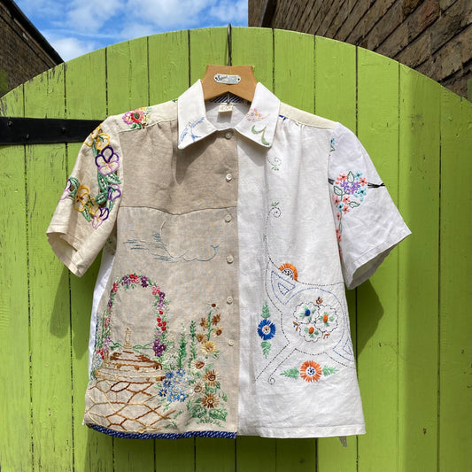Short-sleeved shirt made from vintage hand embroidered tablecloths, mainly white and off-white with colourful embroidery. Photographed hanging on a green gate