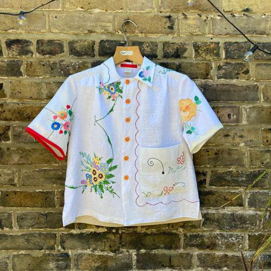 short-sleeved shirt made from recycled tablecloths is mainly white and off-white with colourful embroidered and appliquéd flowers and orange buttons