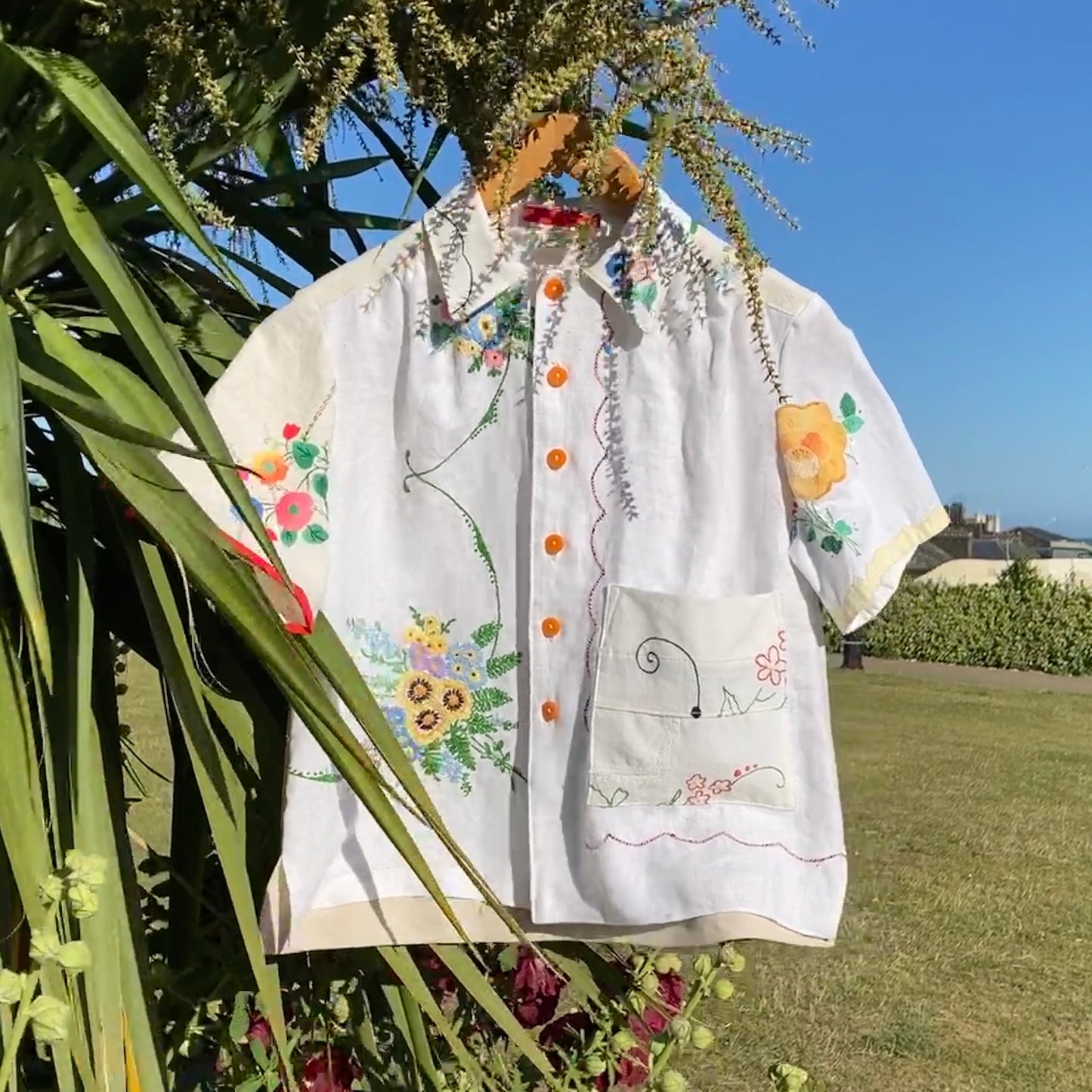 short-sleeved shirt made from recycled tablecloths is mainly white and off-white with colourful embroidered and appliquéd flowers and orange buttons, pictured hanging in a palm tree on the sea front